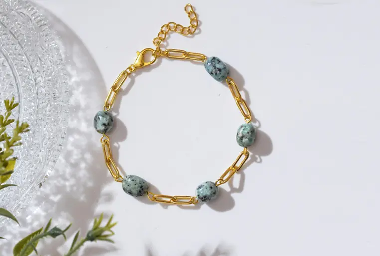 Gold chain bracelet with green and black speckled beads, displayed on a white surface with a leaf and textured glass plate in the background.
