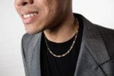 Close-up of a person's lower face wearing a gray blazer, black shirt, and an 18k Saudi Yellow Solid Gold Figaro Chain Unisex necklace. Only the bottom half of the face is visible.