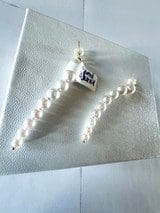 A pair of 18k Gold Akoya Pearls Earrings, one with a white square charm featuring a dark blue design, displayed on a textured white square surface.